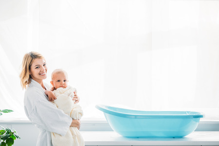 side view of happy mother in bathrobe carrying adorable child covered in towel near plastic baby...