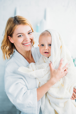 smiling mother in bathrobe holding confused little child covered with towel and looking at cameraの写真素材