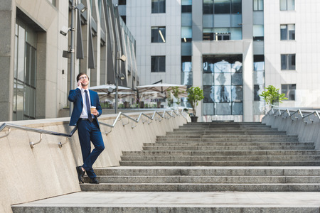 handsome young businessman in stylish suit with coffee to go talking by phone on stairs near business buildingの写真素材