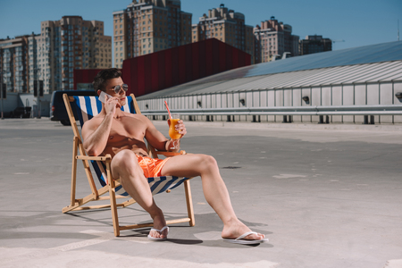 handsome young shirtless man relaxing on sun lounger with cocktail and talking by phone on parkingの写真素材