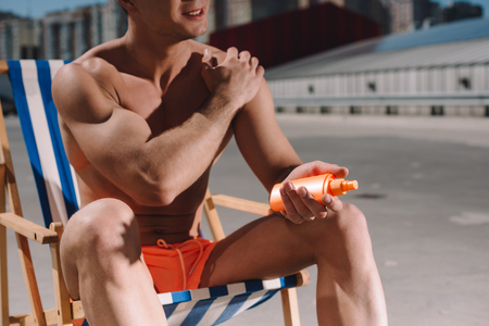 cropped shot of shirtless man sitting on sun lounger and applying sunscreen lotion on parkingの写真素材