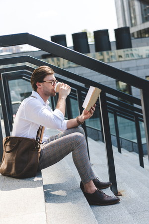handsome young man drinking coffee from paper cup and reading book on stairs on city streetの写真素材