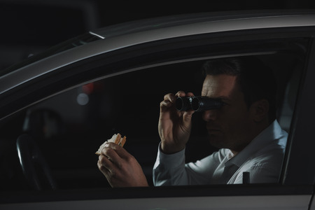 male private detective doing surveillance by binoculars and eating sandwich in carの写真素材