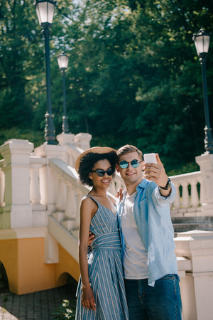 young interracial couple in sunglasses taking selfie on smartphone in parkの写真素材