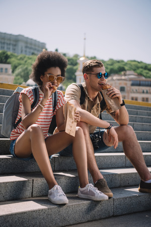 multiethnic couple of travelers sitting on stairs and eating croissantsの写真素材