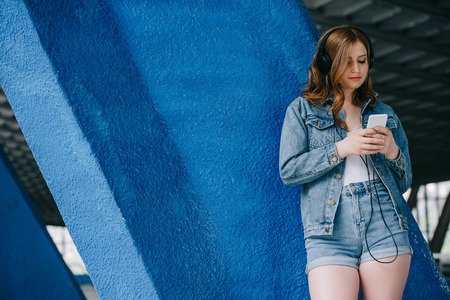 portrait of young fashionable woman in denim clothing and headphones using smartphoneの写真素材