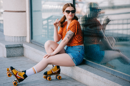 beautiful girl in sunglasses and roller skates smiling at camera while sitting on streetの写真素材