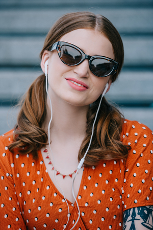 beautiful young woman in sunglasses smiling at camera while listening music in earphones on streetの写真素材