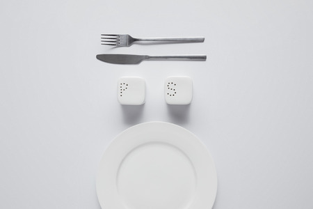 top view of arranged plate, fork, knife, saltcellar and pepper caster on white table, minimalistic conceptの写真素材