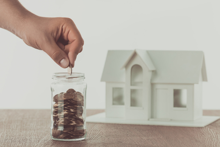 cropped image of man putting coin into glass jar with small house on table, saving conceptの写真素材