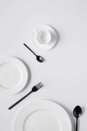 top view of plates, cup, fork and black spoons on white tableの写真素材