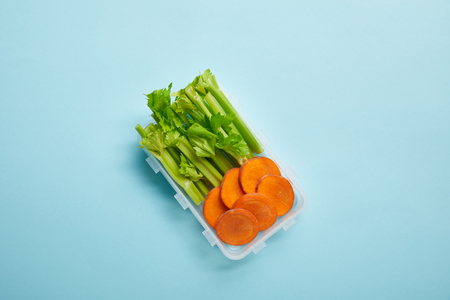 top view of food container full of healthy celery and carrot slices isolated on blueの写真素材