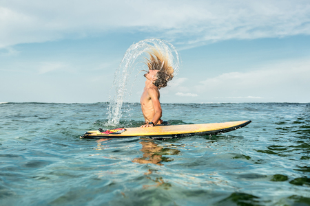 side view of shirtless male surfer emerging from water with surfing board in ocean at Nusa Dua...