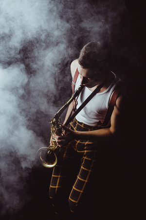 high angle view of stylish young musician playing saxophone in smoke on blackの写真素材