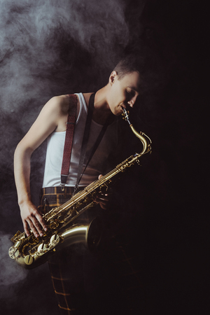 handsome young musician playing saxophone in smoke on blackの写真素材