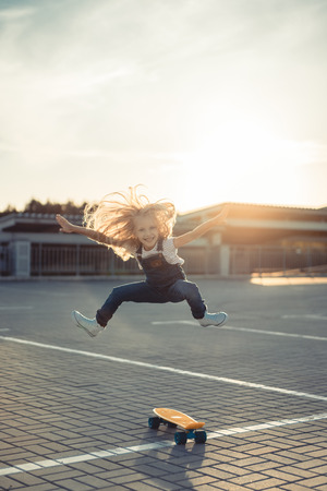 happy little child jumping with wide arms near skateboard at parking lotの写真素材