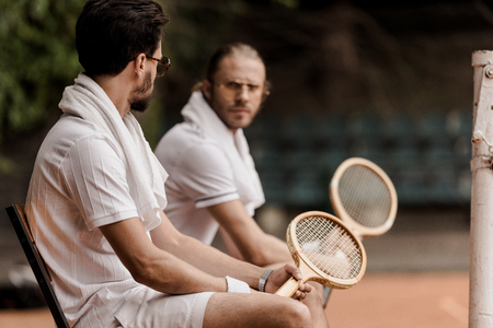 serious retro styled tennis players sitting on chairs with towels and rackets at tennis courtの写真素材