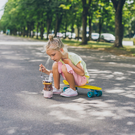 selective focus of little child sitting on skateboard and eating dessert by spoon at urban streetの写真素材