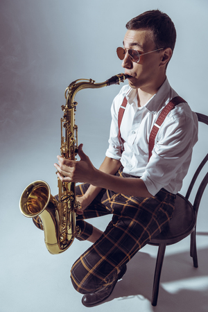 high angle view of stylish young performer sitting on chair and playing saxophone on greyの写真素材