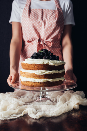 cropped shot of woman holding freshly baked blackberry cake on glass stand on blackの写真素材