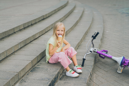 cute child eating ice cream while sitting on city steps near bicycle aloneの写真素材