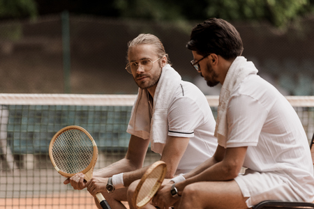 retro styled tennis players sitting on chairs with towels and rackets at tennis courtの写真素材
