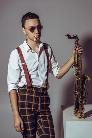 stylish young musician with cigarette holding saxophone on greyの写真素材