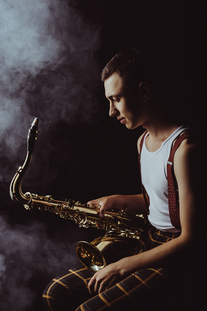 side view of stylish young jazzman sitting and holding saxophone in smoke on blackの写真素材