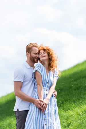 happy redhead man embracing girlfriend from behind on grassy hillの写真素材