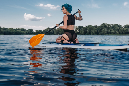 athletic tattooed girl sitting on paddle board on river