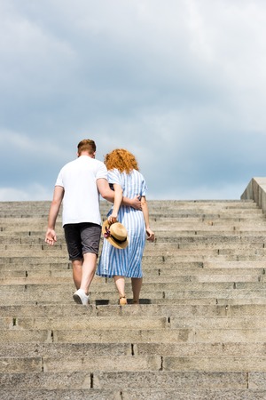 rear view of man embracing redhead girlfriend and walking on stairsの写真素材