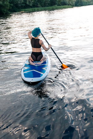 back view of sportive girl with blue hair paddle surfing on river