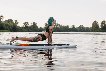tattooed woman with blue hair practicing yoga on paddleboard in water. Upward facing dog pose (Urdhva Mukha Svanasana)の写真素材