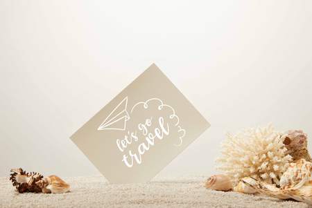 close up view of coral, seashells and blank card on sand on grey background with lets go travel...