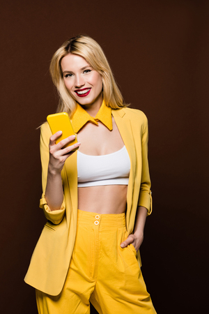 beautiful stylish blonde woman holding yellow smartphone and smiling at camera isolated on brownの写真素材