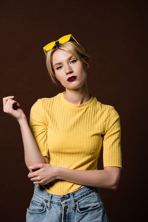 portrait of beautiful stylish blonde girl in yellow sunglasses on head looking at camera isolated on brownの写真素材
