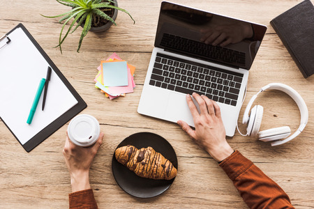 cropped image of man working at table with laptop, headphones, textbook, clipboard, post it, potted plant, coffee cup and croissantの写真素材