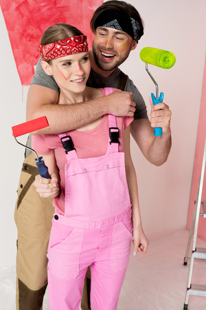 smiling man with paint roller embracing girlfriend in working overall in front of half painted wallの写真素材