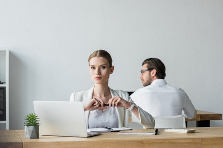 businesswoman sitting at workplace and looking at camera while his colleague working on backgroundの写真素材