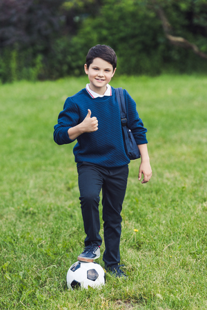 smiling kid with soccer ball and backpack showing thumb up on grass fieldの写真素材