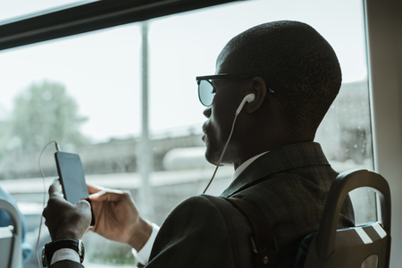 Stylish confident businessman in earphones using smartphone while taking trainの写真素材