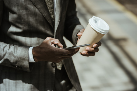 Cropped view of african american businessman wearing suit with smartphone and coffee cupの写真素材