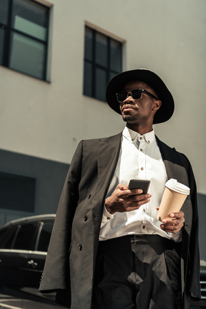 Fashionable african american businessman wearing fedora hat using smartphone and holding coffee cupの写真素材