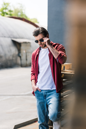 front view of young stylish man standing near wooden pallets and adjusting sunglassesの写真素材