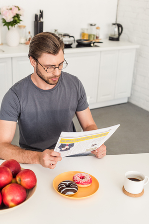 young man in eyeglasses reading newspaper at table with apples, donuts and coffee cupの写真素材