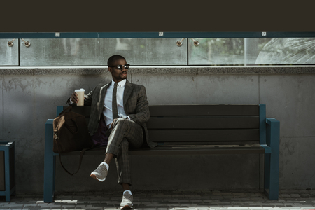 Stylish confident businessman with coffee cup sitting on train station benchの写真素材