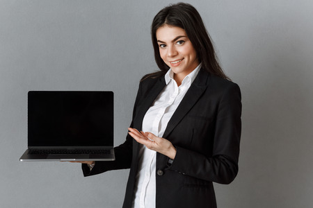 smiling businesswoman pointing at laptop with blank screen against grey wall backgroundの写真素材