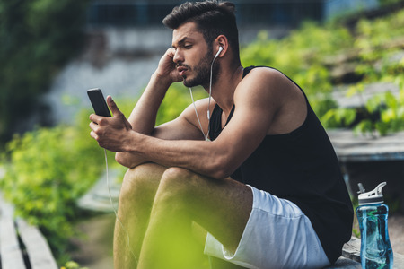 focused sportsman with bottle of water listening music with smartphone and earphones on bench at sport playgroundの写真素材