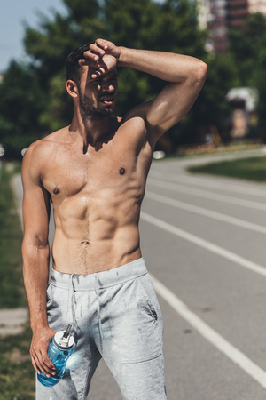 sweat shirtless young man standing on running track after workout and wiping foreheadの写真素材