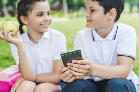 schoolchildren using smartphone together and chatting while sitting on grassの写真素材
